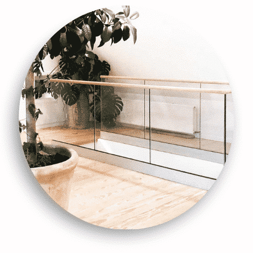 Ein Glasgeländer mit Klammern und einem Handlauf aus Holz, im Innenbereich entlang eines Dachbodens installiert. Das Geländer kombiniert transparentes Glas und warme Holzdetails für eine luftige und sichere Abgrenzung im Raum. Grünflächen und helles Holz Parkett ergänzen die natürliche Atmosphäre.