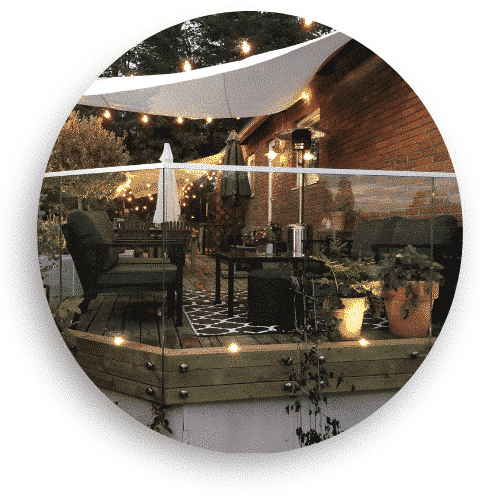 Terrasse mit Ganzglasgeländer auf Holzkonstruktion, umgeben von Gartenmöbeln, Pflanzen und stimmungsvoller Beleuchtung.