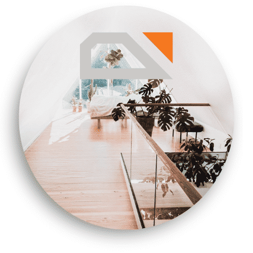 Innenaufnahme, die ein Glasgeländer mit Holzdetails in einem Dachgeschoss zeigt. Das Geländer erstreckt sich entlang des holzverkleideten Bodens und der Blick aus dem Fenster lässt natürliches Licht herein, das einen hellen und luftigen Eindruck vermittelt. Mehrere Pflanzen schmücken den Raum und sorgen für eine harmonische und grüne Atmosphäre.