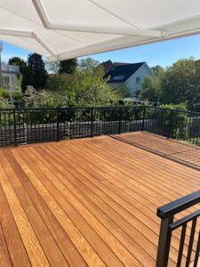 Schmiedeeisernes Geländer in Schwarz um Holzterrasse mit Markise, sonnenbeschienenen Dielen und grüner Umgebung.