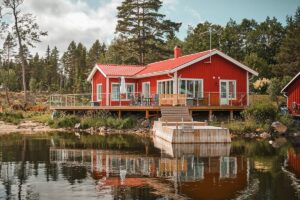 Terrasse mit Glasgeländer rund um rotes Seehaus, Treppe zum Steg und spiegelglatter Wasserfläche im Vordergrund.
