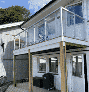 Glasgeländer mit weißen, eckigen Aluminiumpfosten auf Balkon mit Holzdielen und Fassade in Holzoptik.