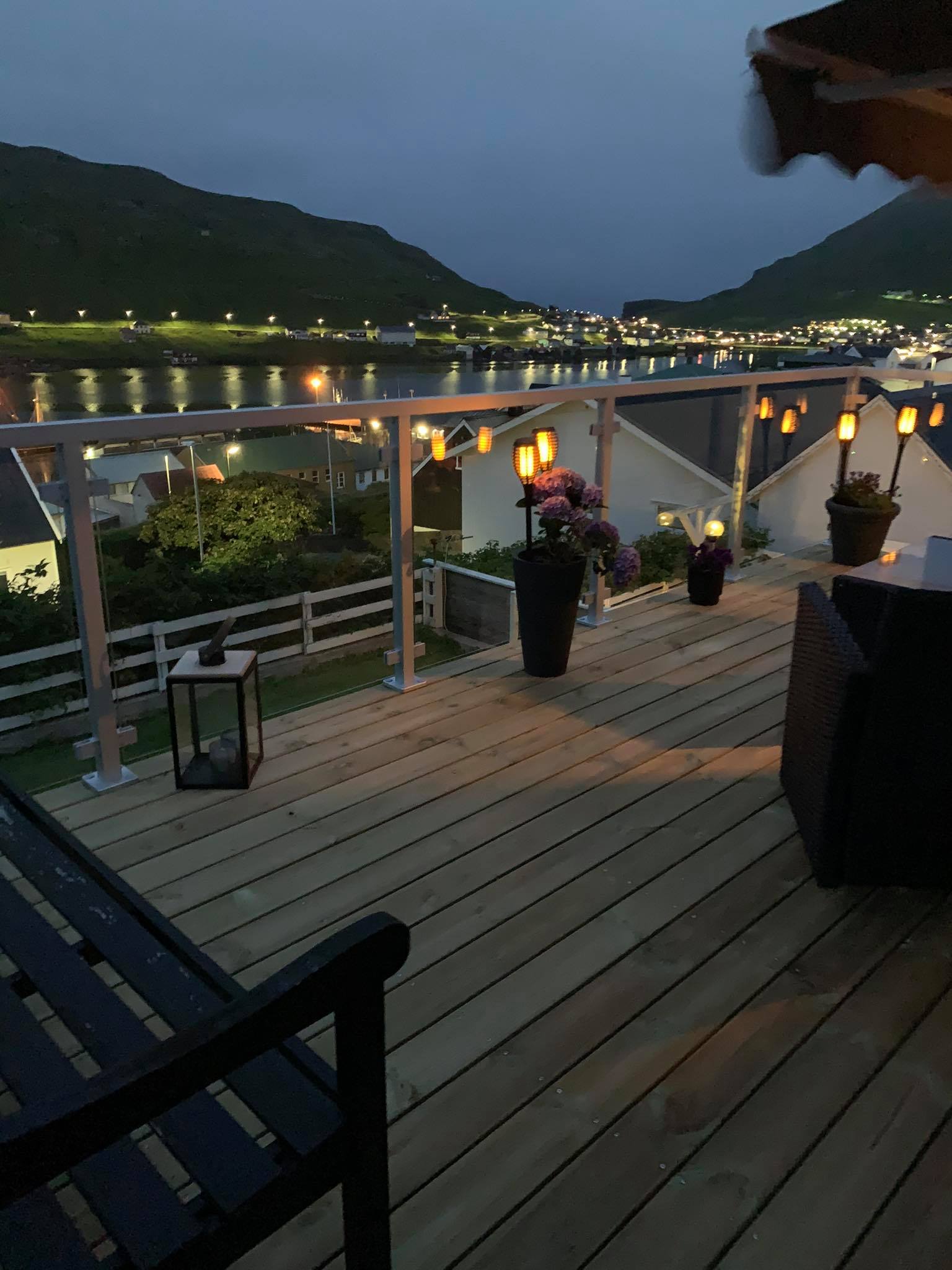 Weißes Aluminiumgeländer mit Klarglas rund um beleuchtete Terrasse mit Ausblick auf Fjord und Berge in der Dämmerung.