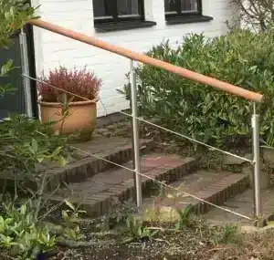 Treppengeländer aus Edelstahl mit Drähten und einem Handlauf aus Holz. Installiert entlang einer Backsteintreppe, die zu einem Haus mit weißer Fassade und schwarzen Fenstern führt. Umgeben von Pflanzen und einer Topfpflanze am Eingang.