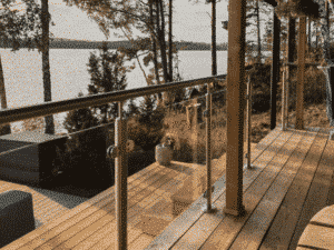 Glasgeländer mit Edelstahlpfosten und Handlauf aus Holz montiert auf einem Holzbalkon. Das Geländer hat Klammern, die die Glasscheiben an Ort und Stelle halten. Im Hintergrund sieht man einen von Wald umgebenen See sowie eine absteigende Holztreppe, die zum Wasser führt.