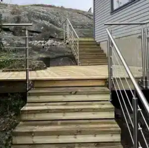 Treppe und Balkon aus druckimprägniertes Holz mit Edelstahlgeländer Das Geländer hat horizontale Fullstäbe und wird entlang von Treppen und Podesten montiert. Das Haus hat eine Fassade aus grauen Paneelen und Bergen im Hintergrund.