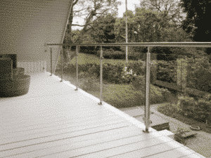 Glasgeländer mit Edelstahlpfosten und Handläufen auf einem weiß gestrichenen Balkon mit Blick auf einen üppigen Garten.