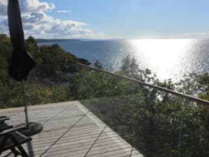 Stilvolles, pfostenloses Glasgeländer mit Holzhandläufen auf einem Balkon am Meer, das einen freien Blick auf das Wasser und den Horizont ermöglicht.