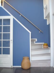 Treppe mit weiß lackierten Stufen und einem an der Wand montierten Handlauf aus Edelstahl. Der Handlauf wird mit dezenten Wandhalterungen an einer blauen Wand befestigt. Im Raum sind eine weiße Glastür und dekorative Tontöpfe zu sehen.