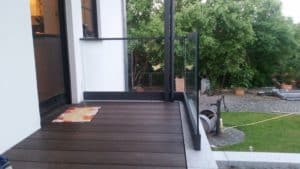Glasgeländer mit schmalem Tor auf dunklem Holzdeck am Eingang, mit schwarzem Aluminiumrahmen und Blick auf Garten und gepflasterten Sitzbereich.