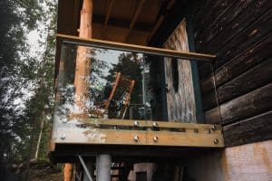 Pfostenfreies Glasgeländer auf kleinem Holzbalkon, punktgefasst mit Holzhandlauf, montiert an Blockhaus in waldreicher Umgebung.