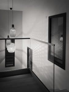 Moderne Innenarchitektur mit pfostenfreiem Glasgeländer und Hängelampen, die grafische Lichtmuster auf Wände mit Holzfußboden werfen.