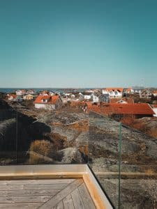 Nahaufnahme einer Ecke eines pfostenfreien Glasgeländers mit Klarglas und Holzrahmen, montiert auf Terrasse mit Blick auf Felsen und Küstenlandschaft.