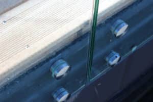 Nahaufnahme einer Punkthalterung eines pfostenfreien Glasgeländers mit Edelstahlabstandshaltern, montiert auf Holz- und Betonuntergrund.