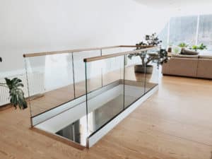 Elegantes pfostenfreies Glasgeländer rund um Treppenöffnung in moderner Dachwohnung mit Holzhandlauf, weißer Sockelleiste und hellem Parkett.
