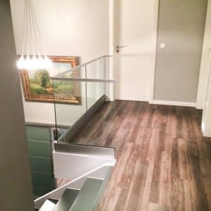 Pfostenfreies Glasgeländer im oberen Treppenbereich mit weißem Handlauf und Klarglas, montiert auf dunklem Holzboden in modernem Innenraum.