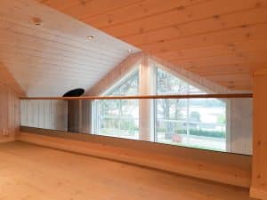 Pfostenfreies Glasgeländer im Innenbereich vor großem Dreiecksfenster, mit Holzverkleidung an Decke und Wänden und Ausblick ins Grüne.