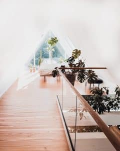 Helle und stilvolle Dachwohnung mit pfostenfreiem Glasgeländer und Holzhandlauf, eingerichtet mit Pflanzen, Holzfußboden und Bett am großen Dreiecksfenster.
