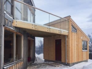 Pfostenfreies Glasgeländer auf Holz-Balkon in Berglandschaft, mit Holzbefestigung und Holzhandlauf zwischen zwei Holzhäusern im Winter.
