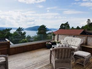 Stilvolles pfostenfreies Glasgeländer auf holzverkleideter Terrasse mit Blick auf Wald und Wasser, ergänzt durch Lounge-Gruppe und Blumentöpfe.