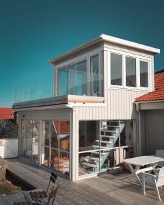 Modernes, pfostenfreies Glasgeländer mit Klarglas und Holzsockel, montiert auf einem weißen Haus mit Meeresnähe.