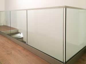 Pfostenfreies Glasgeländer vor großem Dreiecksfenster, mit weißer Holzverkleidung und Holzhandlauf – für helle, harmonische Innenarchitektur.